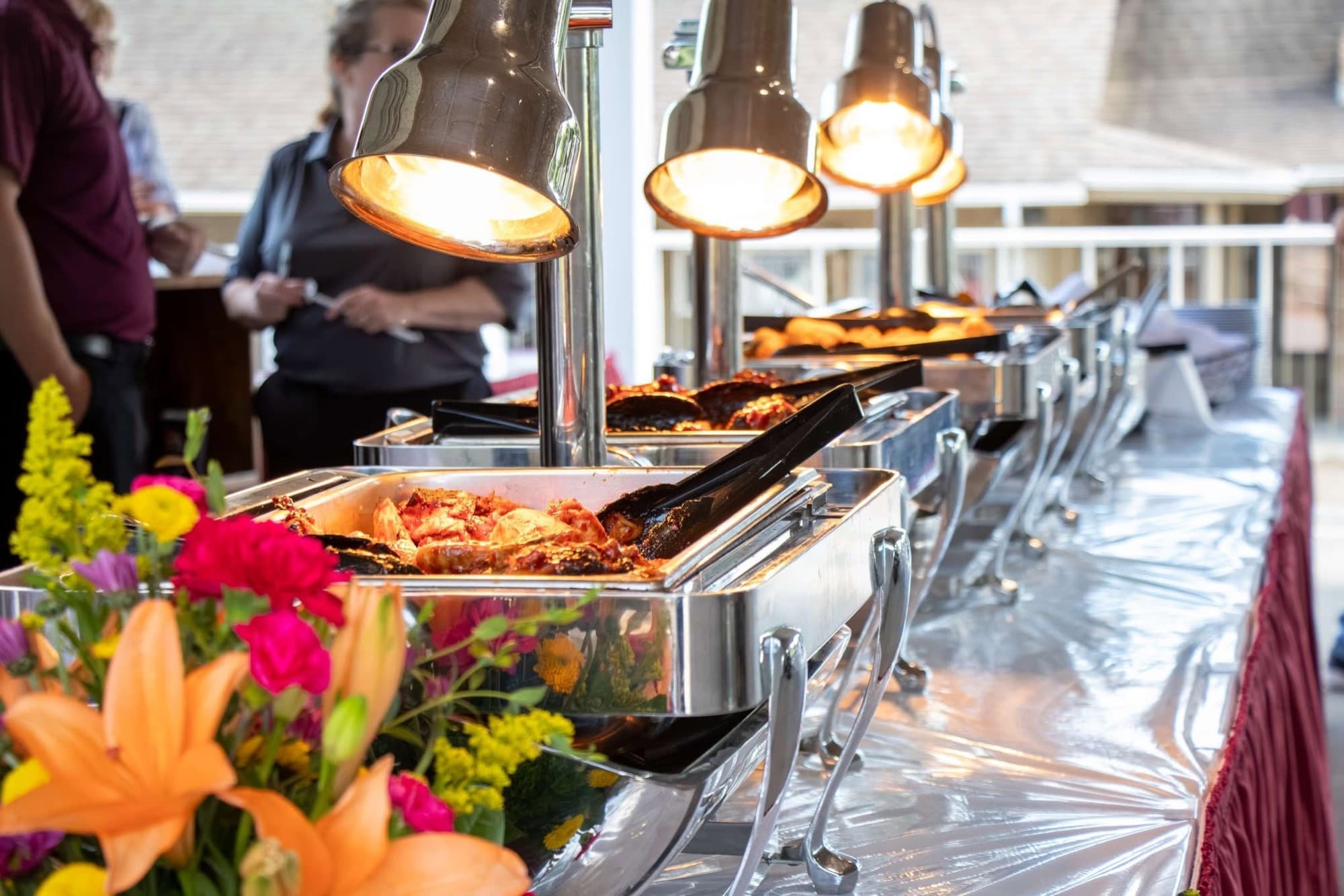 Buffet line at a Fortress event, chafing dishes under heat lamps with fresh flowers, staff serving in the background