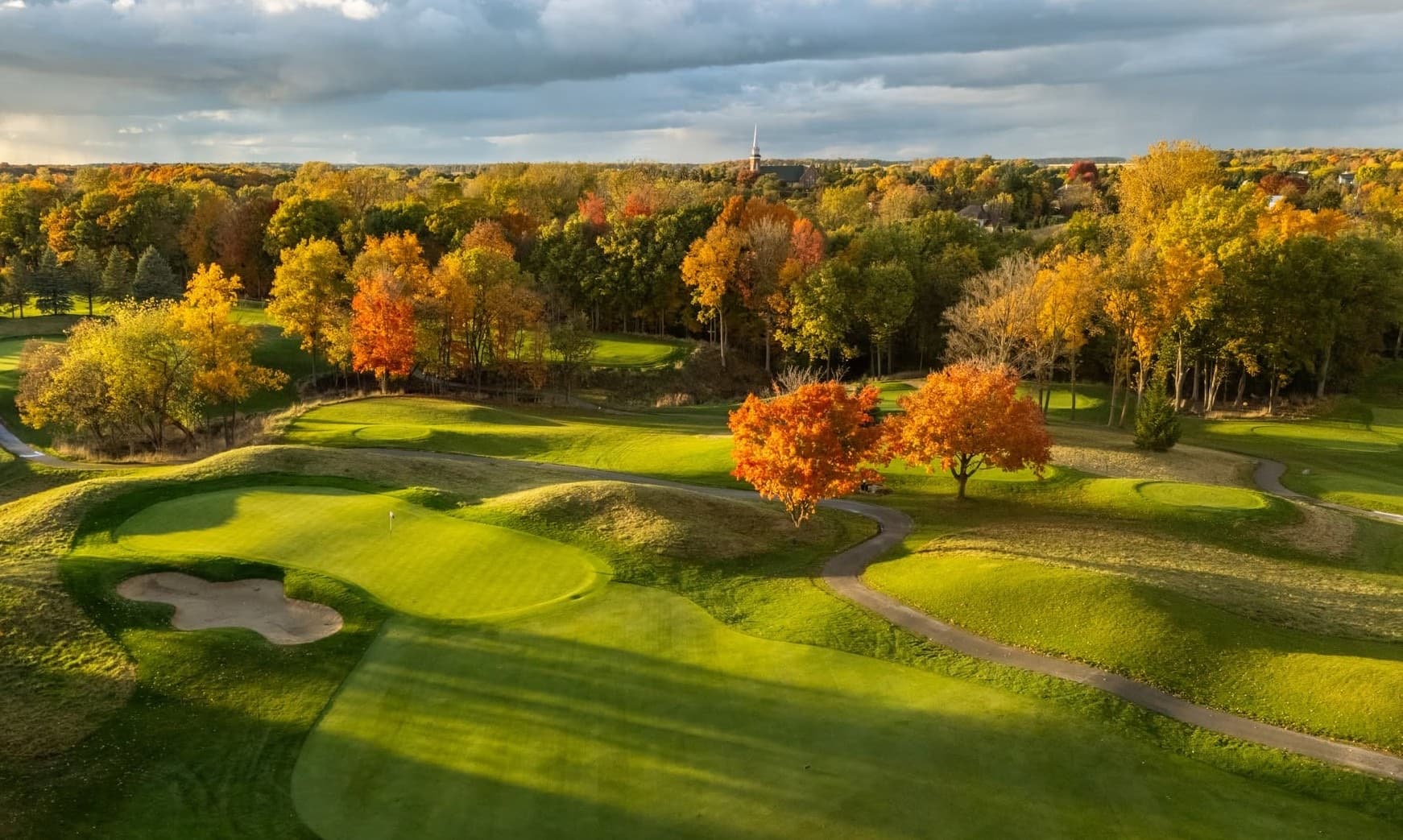 Aerial of the 1st green at The Fortress with the fairway sweeping toward the Frankenmuth steeple in autumn